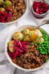 A white bowl filled with savoury minced beef in gravy with boiled new potatoes, peas, and red cabbage, topped with fresh parsley. There is a fork sticking out of the bowl. The bowl is on a white background next to a small dish of red cabbage There is a further bowl just peeking into shot.