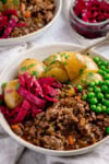 A white bowl filled with savoury minced beef in gravy with boiled new potatoes, peas, and red cabbage, topped with fresh parsley. The bowl is on a white background next to a small dish of red cabbage There is a further bowl just peeking into shot.