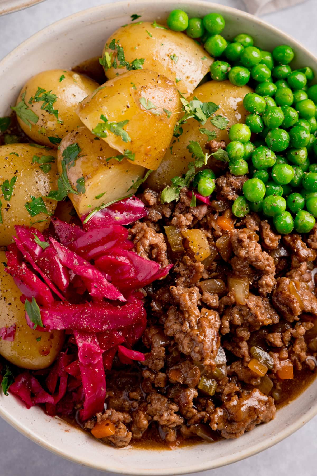Minced beef in gravy in a white bowl with boiled potatoes, green peas, and pickled red cabbage, all garnished with chopped parsley.