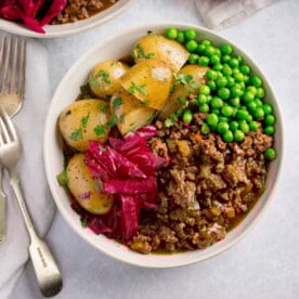 A bowl of savoury minced beef in gravy with, boiled new potatoes, peas, and red cabbage, topped with fresh parsley. The bowl is on a white background next to two silver forks. There is a further bowl just peeking into shot.