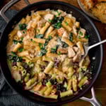 A frying pan with creamy rigatoni alfredo with chicken and kale, and grated cheese, with a spoon resting in the dish.