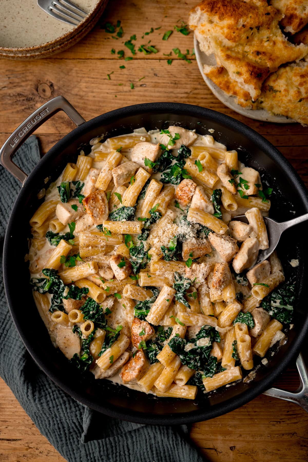 A frying pan filled with creamy rigatoni alfredo pasta, chicken pieces, and kale, topped with grated cheese and parsley. Garlic bread is on a plate nearby.