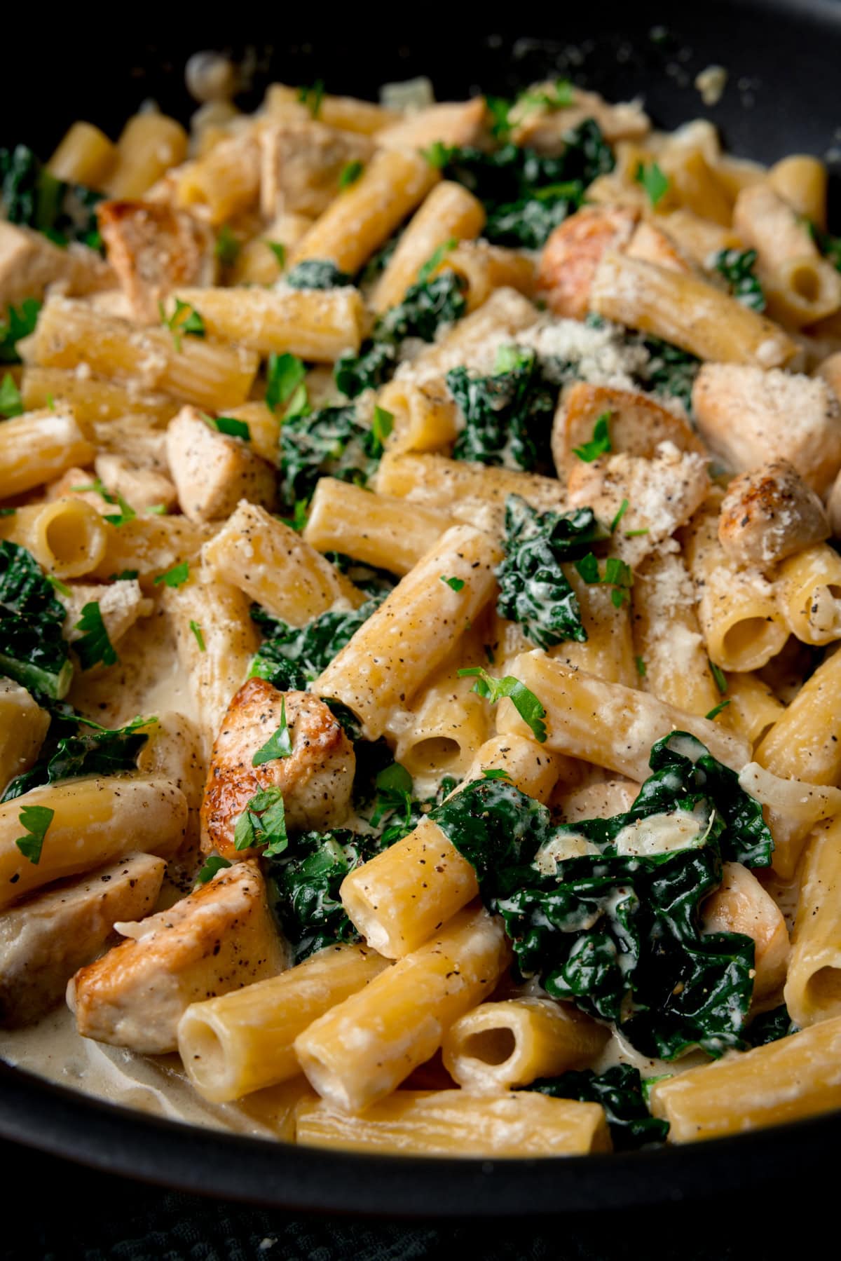 A close up picture of Rigatoni alfredo with chicken, kale, and a creamy sauce, topped with grated cheese and chopped parsley in a black pan.