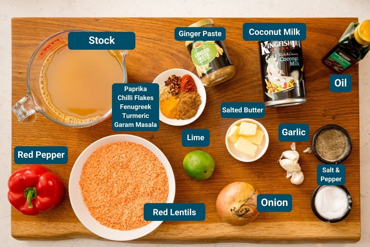 A wooden surface with labelled ingredients for a red lentil dhal, including stock, red pepper, red lentils, spices, lime, onion, garlic, butter, ginger paste, coconut milk, oil, and salt & pepper.