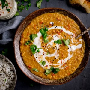 A bowl of red lentil dahl garnished with cream, coriander, and spices, with a side of rice and naan on a dark surface.