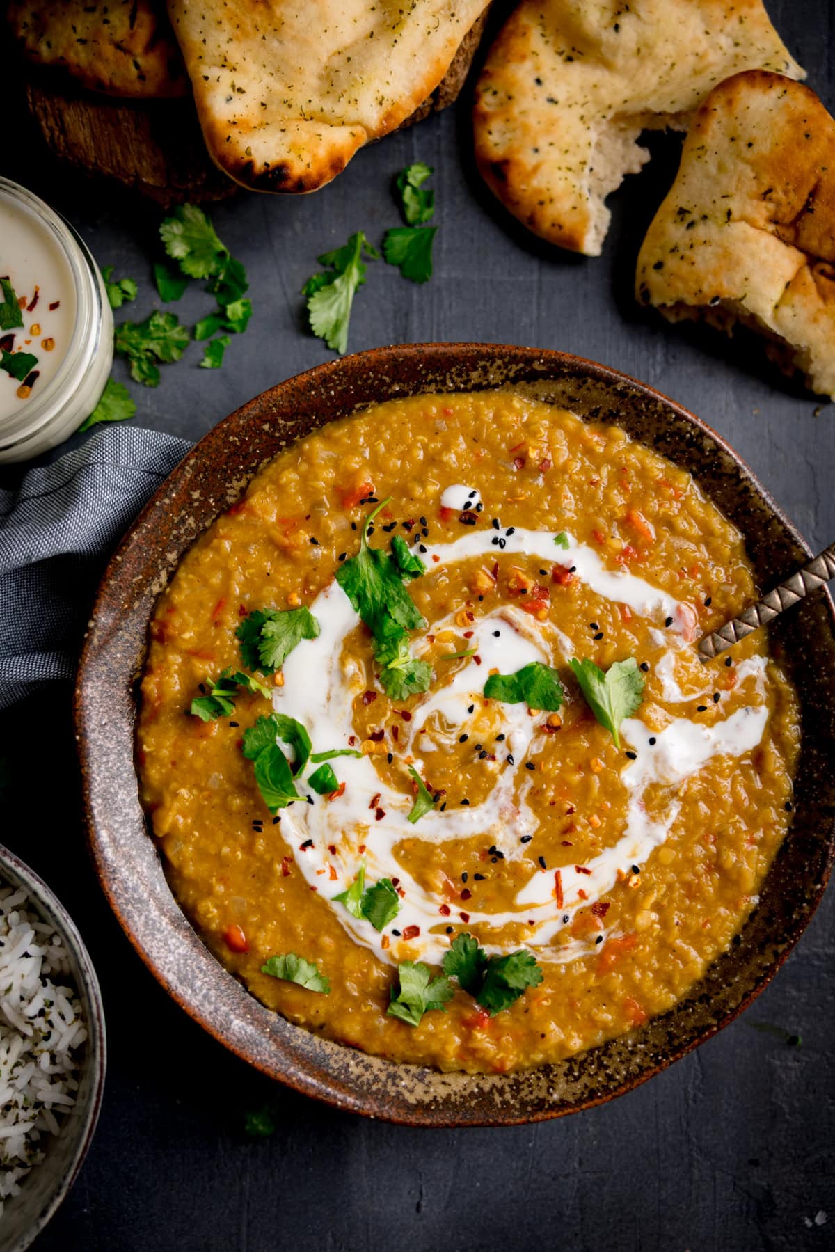 A bowl of lentil dal garnished with cream, coriander, and spices, served with naan bread, rice, and extra coriander on a dark surface.