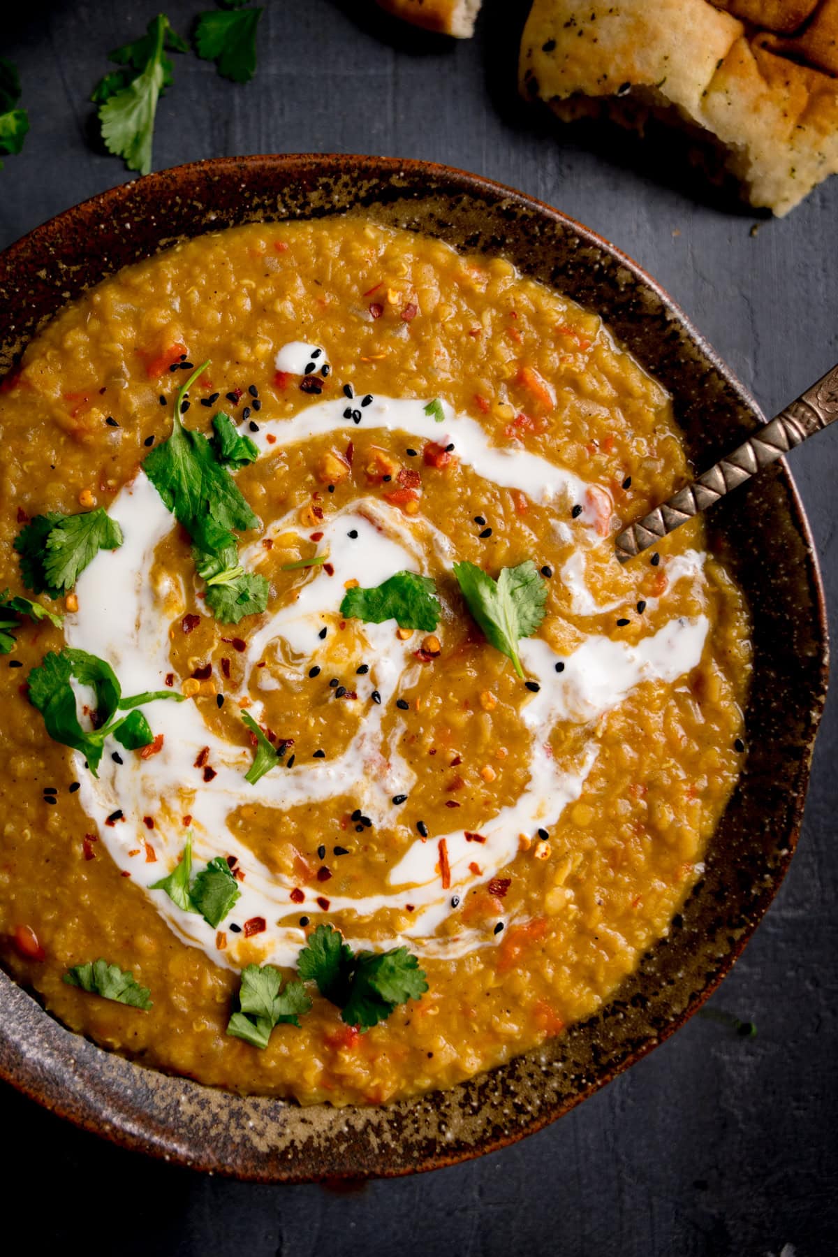 A bowl of lentil curry garnished with a swirl of cream, fresh coriander, and spices, with a piece of bread in the background.