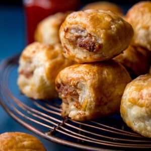 Several golden-brown sausage rolls stacked on a wire cooling rack, with a blurred bottle of sauce in the background.