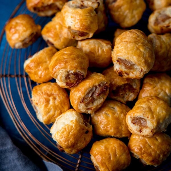 A pile of golden-brown sausage rolls rests on a wire cooling tray, showing flaky pastry and browned meat filling.