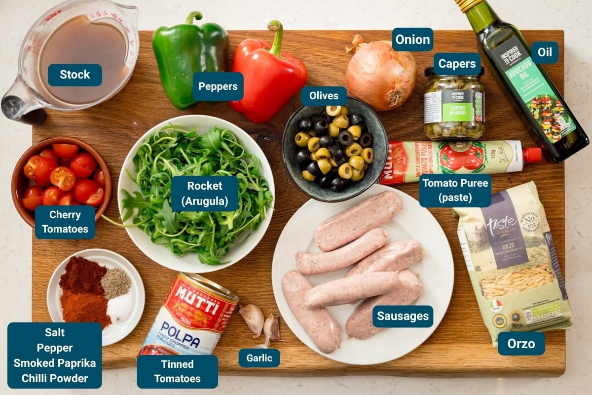 A wooden board displays labelled ingredients for a meal, including sausages, orzo, peppers, rocket, cherry tomatoes, stock, spices, oils, onion, capers, olives, tomato purée, and garlic.
