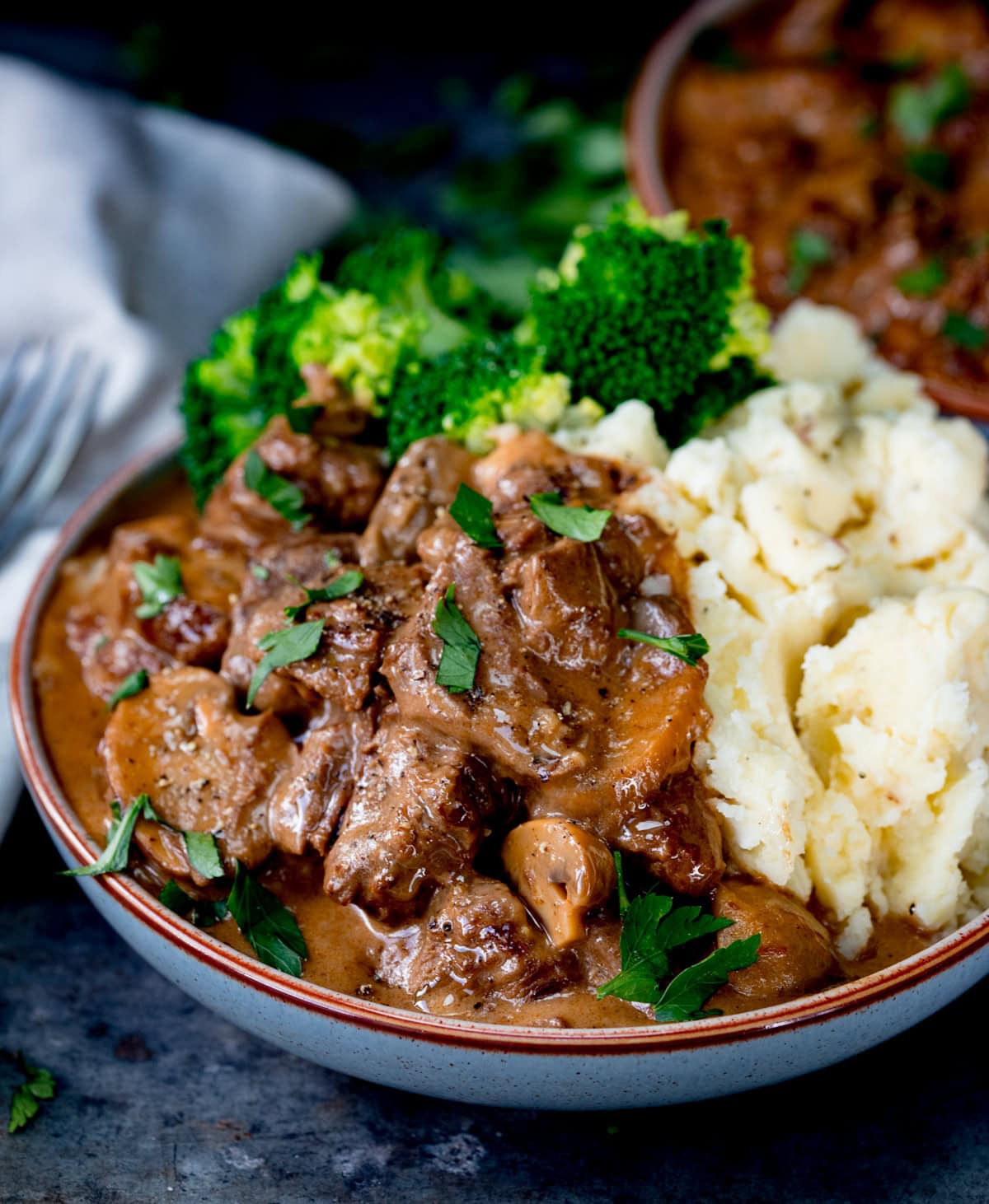 A bowl of slow cooked steak casserole with rich brown gravy, served with mashed potato and steamed broccoli, topped with chopped parsley.