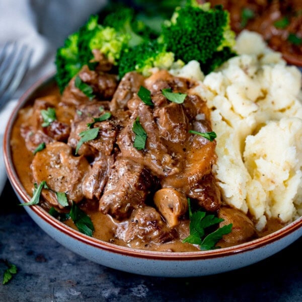 A shallow bowl filled with slow cooked steak diane with mushrooms and rich gravy, served with mashed potato and steamed broccoli, garnished with chopped parsley.