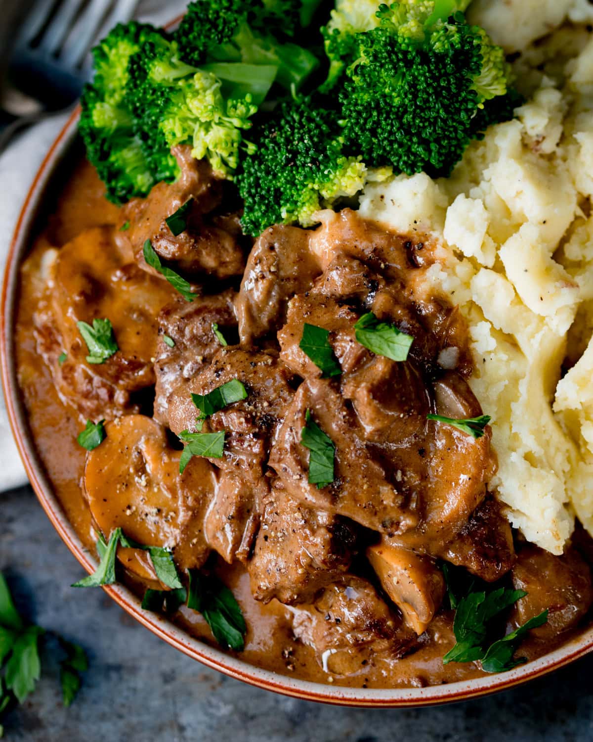 Overhead image of a bowl of slow-cooked steak casserole with mushrooms and gravy, served with mashed potato and steamed broccoli, garnished with chopped parsley.