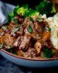 A bowl of slow cooked steak diane casserole with mushrooms in gravy, served with mashed potato and steamed broccoli, topped with chopped parsley.