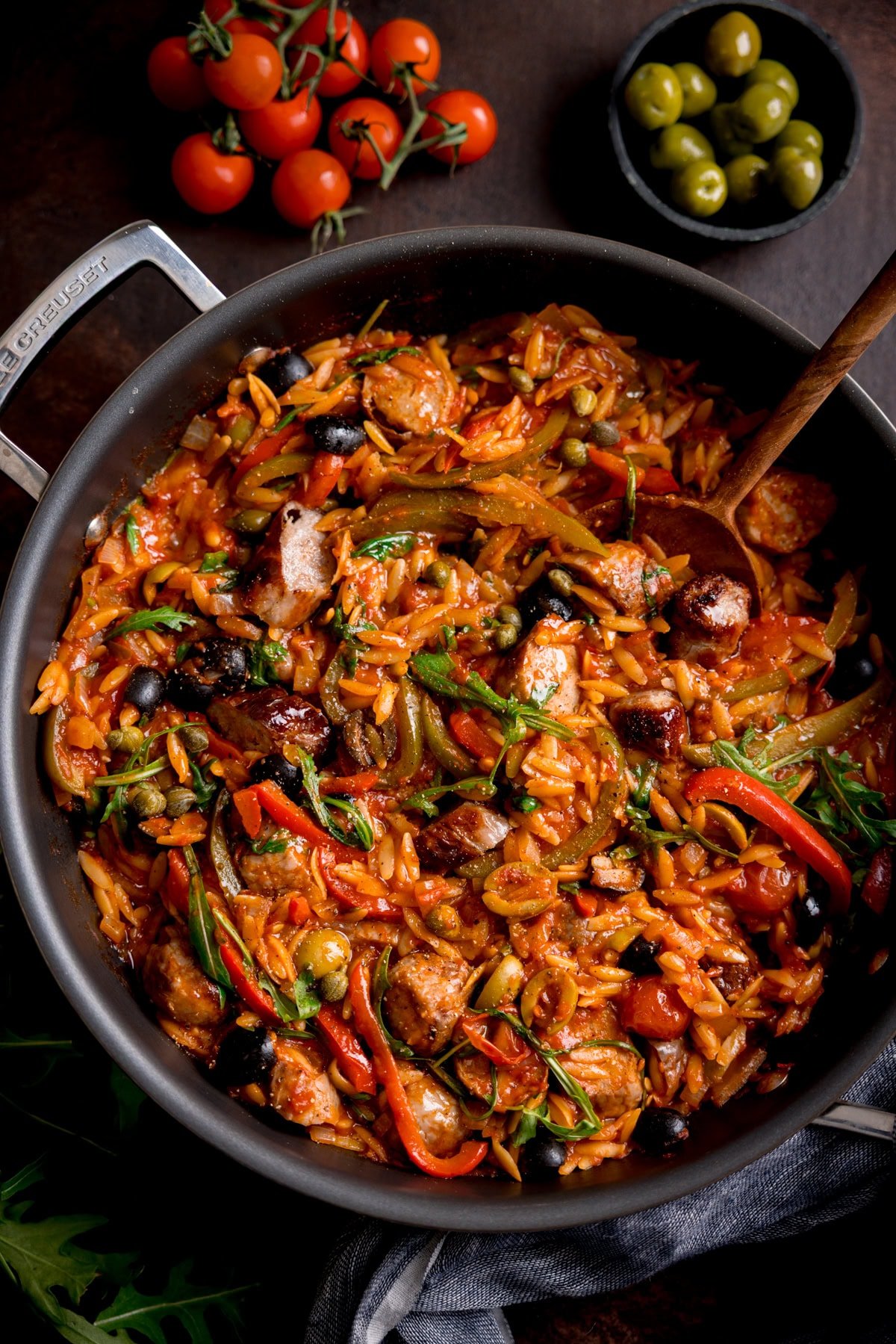A frying pan filled with tomato sausage orzo, vegetables, olives, with a wooden spoon; cherry tomatoes and a bowl of green olives in the background.