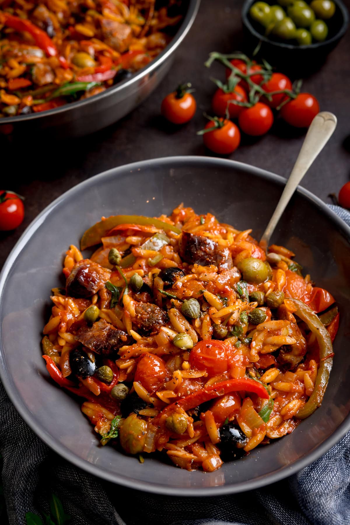 A bowl of tomato sausage orzo pasta with tomato sauce, sliced sausage, capers, olives, cherry tomatoes, and herbs, with a fork at the side.