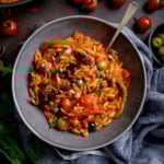 A grey bowl of tomato sausage orzo pasta with tomato sauce, pieces of sausage, peppers, olives, capers, and cherry tomatoes, served with a fork.