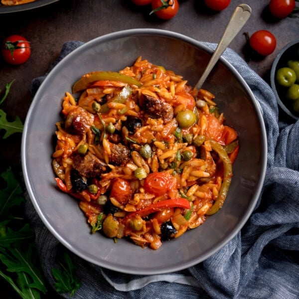 A grey bowl of tomato sausage orzo pasta with tomato sauce, pieces of sausage, peppers, olives, capers, and cherry tomatoes, served with a fork.