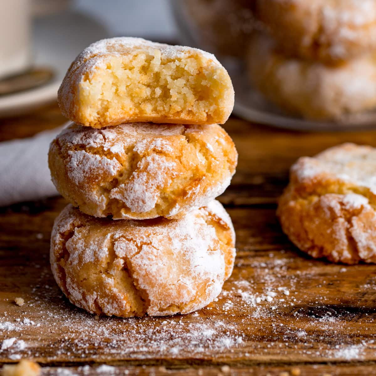 Three powdered amaretti cookies are stacked on a wooden surface, with the top biscuit showing a bite taken out. Crumbs lie nearby.