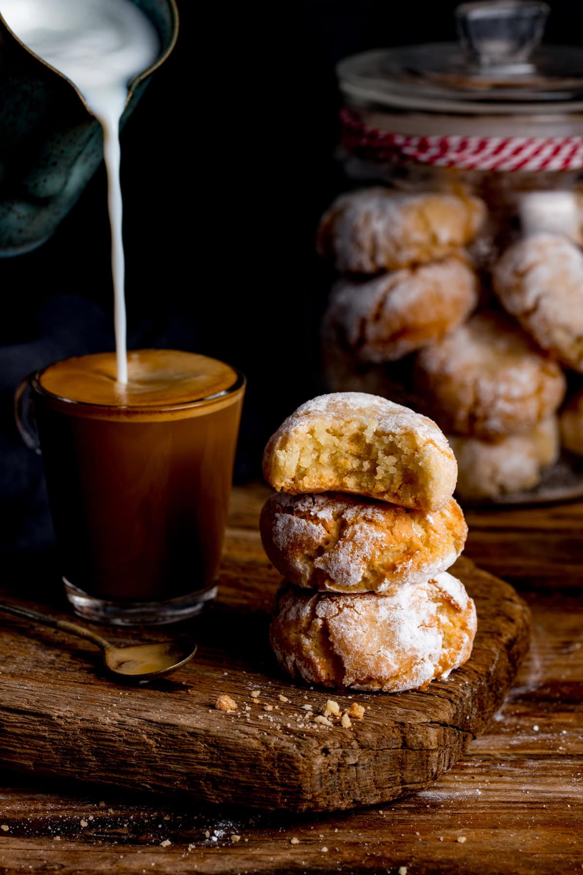 A stream of milk pours into a glass of coffee beside a stack of amaretti cookies, with a jar of more cookies in the background.