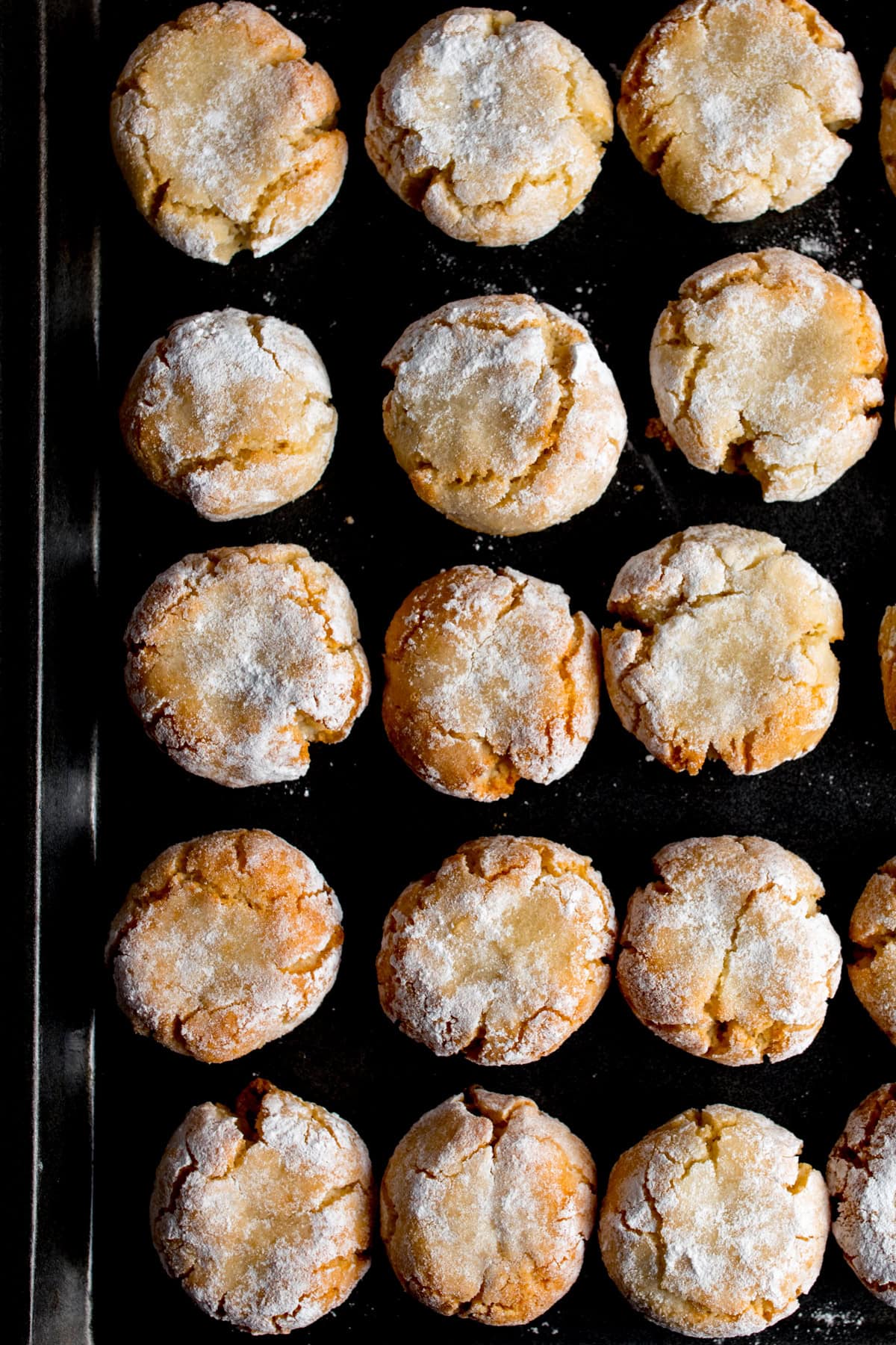 Rows of round, cracked amaretti cookies dusted with icing sugar arranged on a dark baking tray.