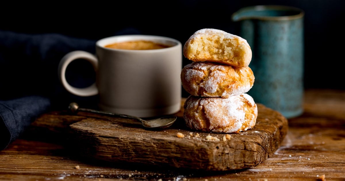A stack of three icing sugar-covered amaretti biscuits sits on a wooden board next to a cup of coffee, a spoon, and a blue-green jug.
