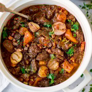 A white pot filled with beef bourguignon containing chunks of beef, carrots, pearl onions, and garnished with parsley. A serving spoon rests inside the pot.