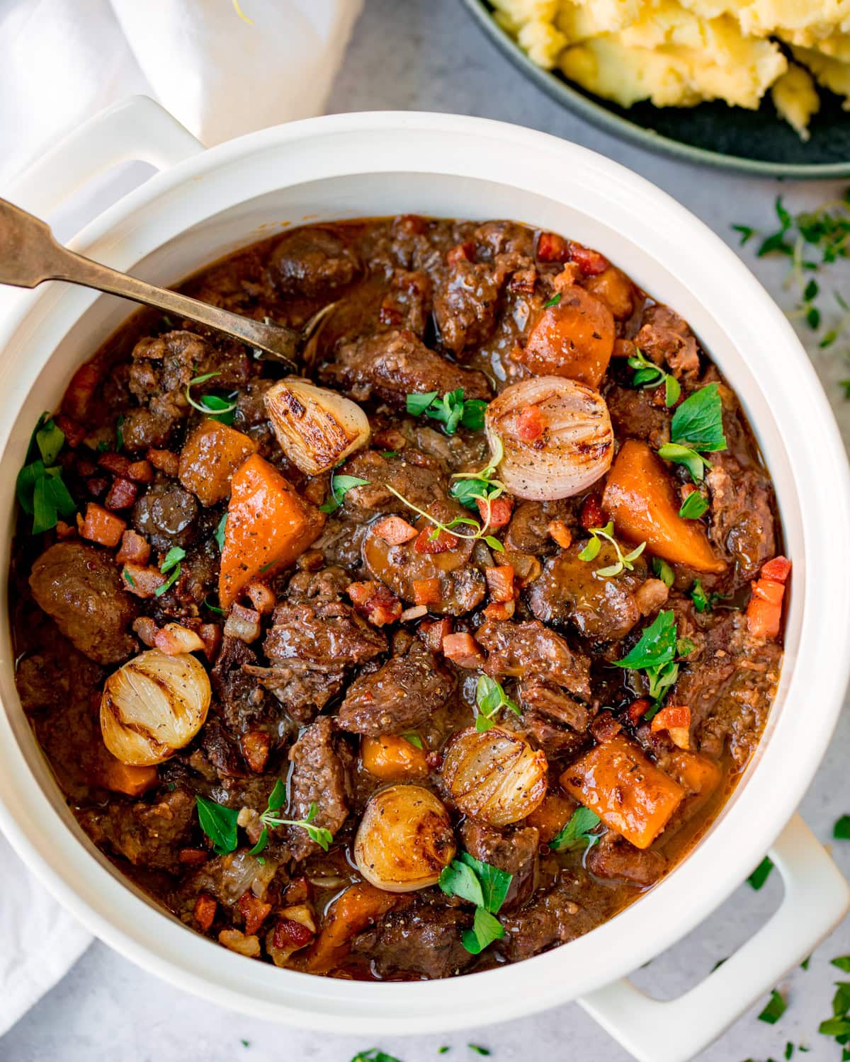 A white pot filled with beef bourguignon containing chunks of beef, carrots, onions, and garnished with fresh herbs.