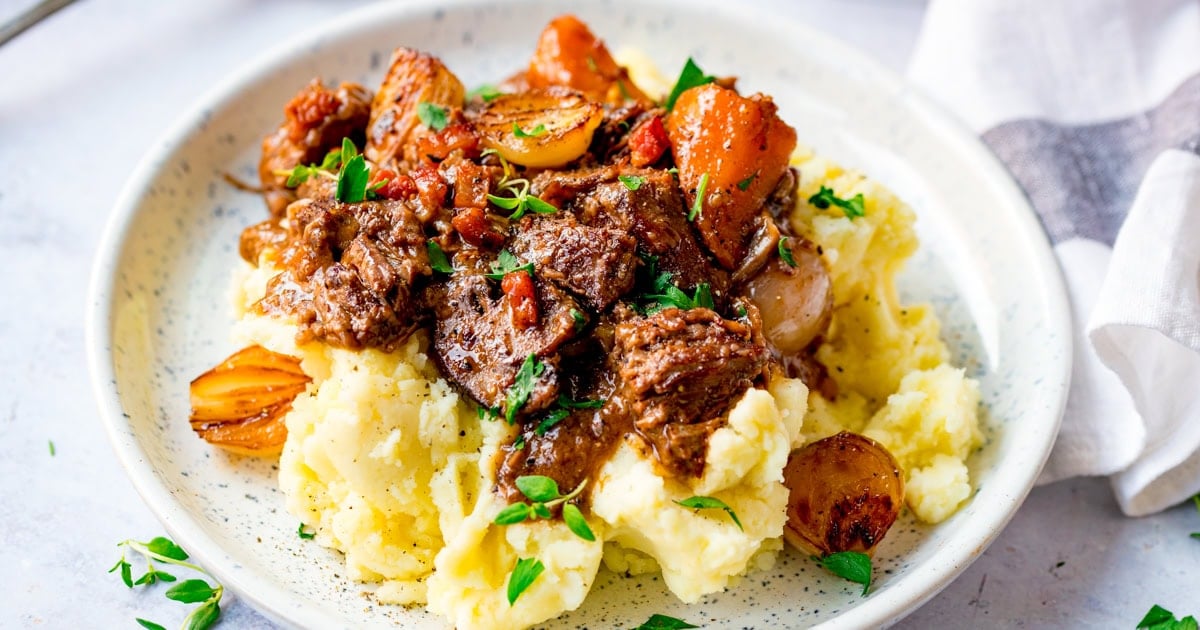 A plate of mashed potato topped with beef bourguignon with carrots, onions, and garnished with chopped parsley.