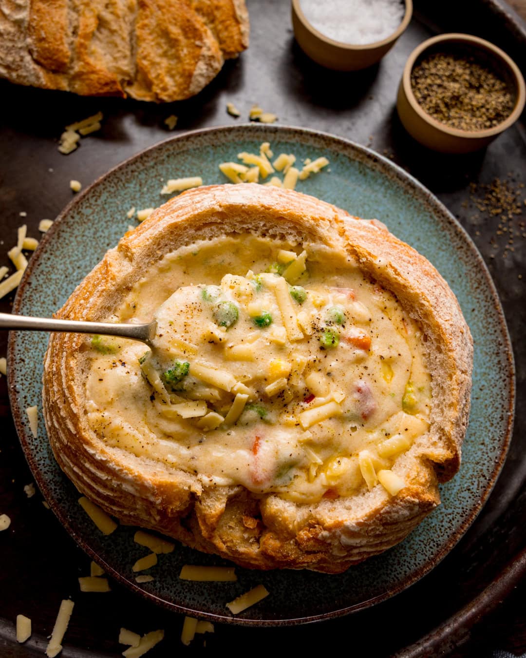 A bread bowl filled with creamy cheddar vegetable soup, topped with grated cheese, served on a plate with a spoon and seasoning dishes nearby.