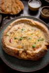A bread bowl filled with creamy Cheddar vegetable chowder, topped with grated cheese and black pepper, served on a blue plate. Two little dishes holding salt and pepper are next to the plate, alongside the top piece of the bread bowl.