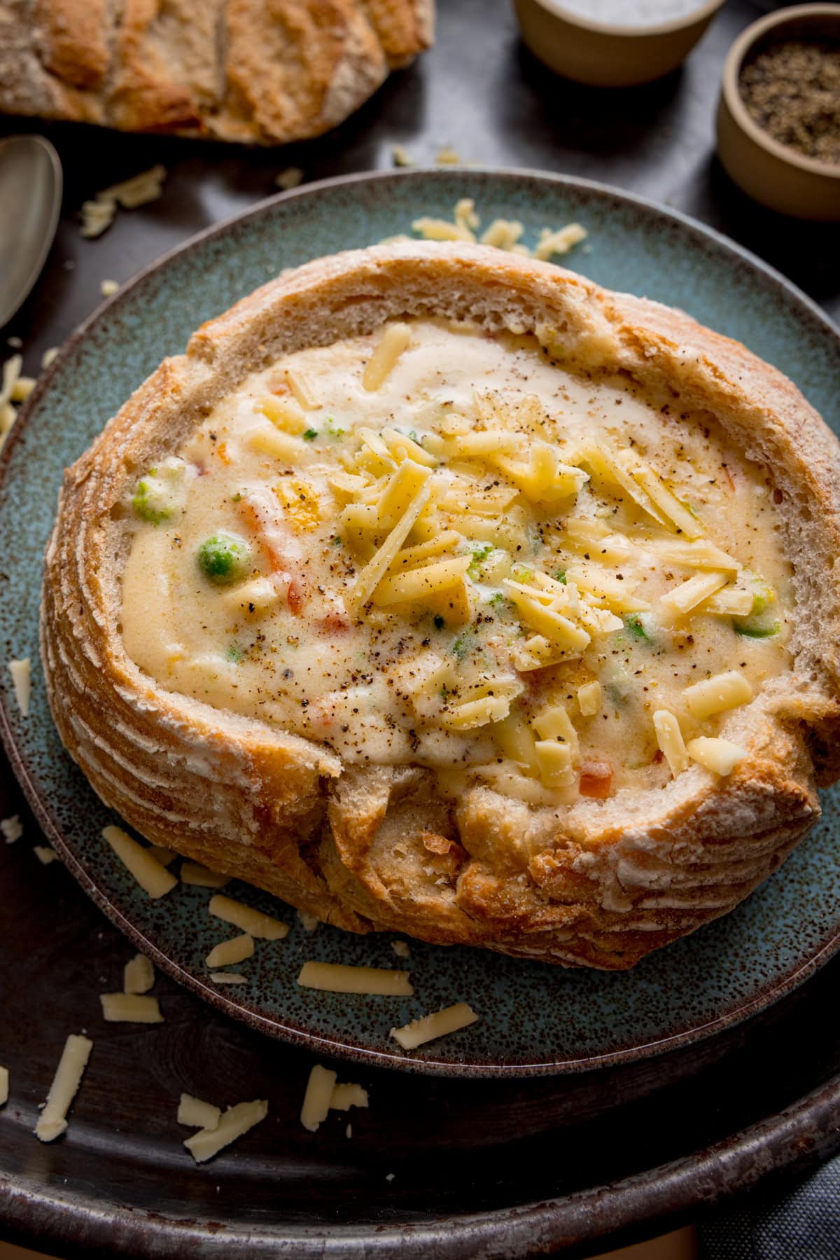 Tall overhead image of a bread bowl filled with creamy vegetable chowder, topped with grated cheddar and ground pepper, on a blue plate with scattered cheese pieces around.