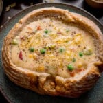 A bread bowl filled with creamy vegetable chowder, topped with grated cheddar cheese and black pepper, served on a blue-green plate.