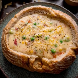 A bread bowl filled with creamy vegetable chowder, topped with grated cheddar cheese and black pepper, served on a blue-green plate.