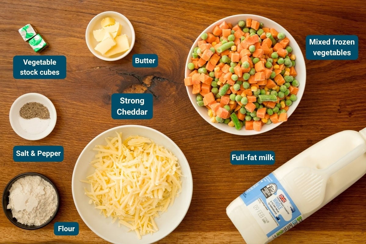 Ingredients for a cheese and Vegetable Chowder laid out on a wooden surface: mixed frozen vegetables, mature cheddar, whole milk, flour, butter, vegetable stock cubes, salt, and pepper.