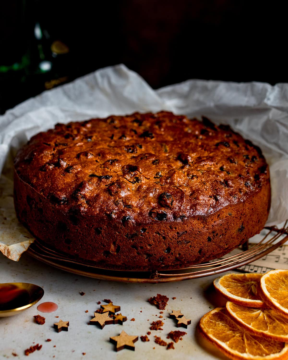 A round fruitcake with a golden-brown crust sits on baking parchment, with dried orange slices and star-shaped decorations nearby.