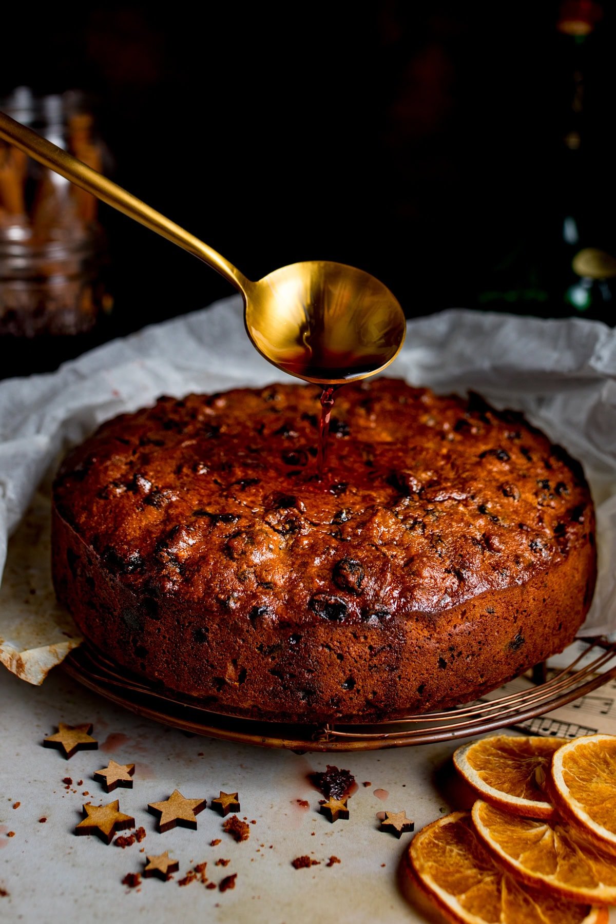 A golden spoon drizzles liquid onto a christmas cake resting on baking parchment, with dried orange slices and star-shaped decorations nearby.