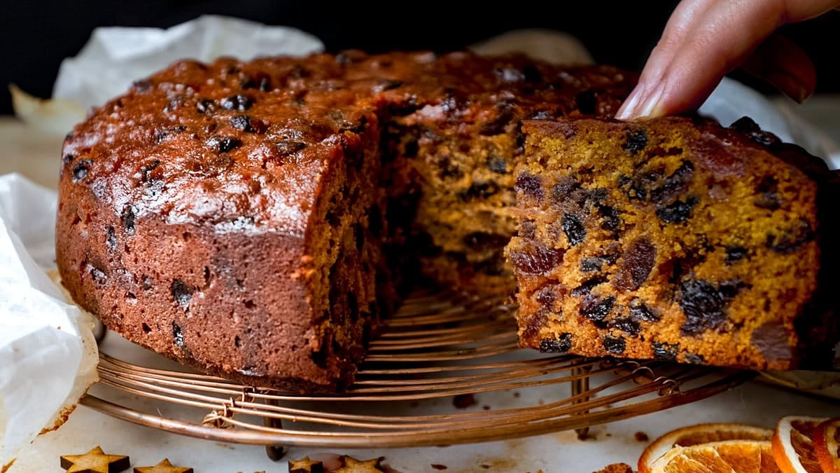 A close-up of a fruitcake on a wire rack, with a hand lifting a slice to show the dense, fruit-filled centre.