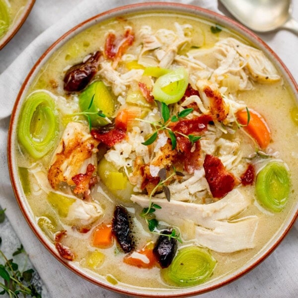 A bowl of cock-a-leekie soup with sliced leeks, carrots, shredded chicken, bacon pieces, herbs and black olives sits on a white cloth with a spoon nearby.