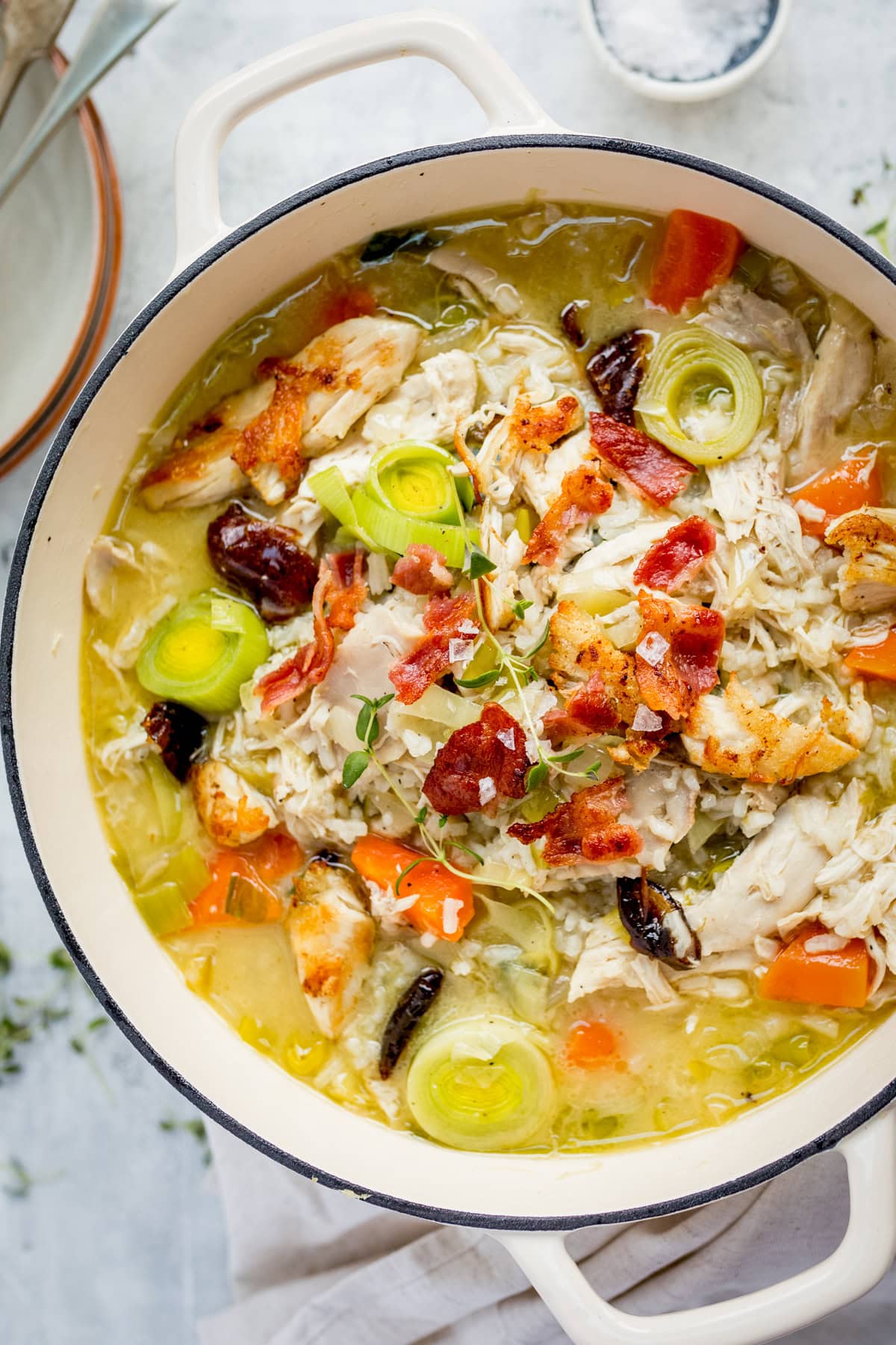 A pot of cock-a-leekie soup with chicken, rice, carrots, leeks, shredded chicken, bacon pieces, and herbs in a light broth.