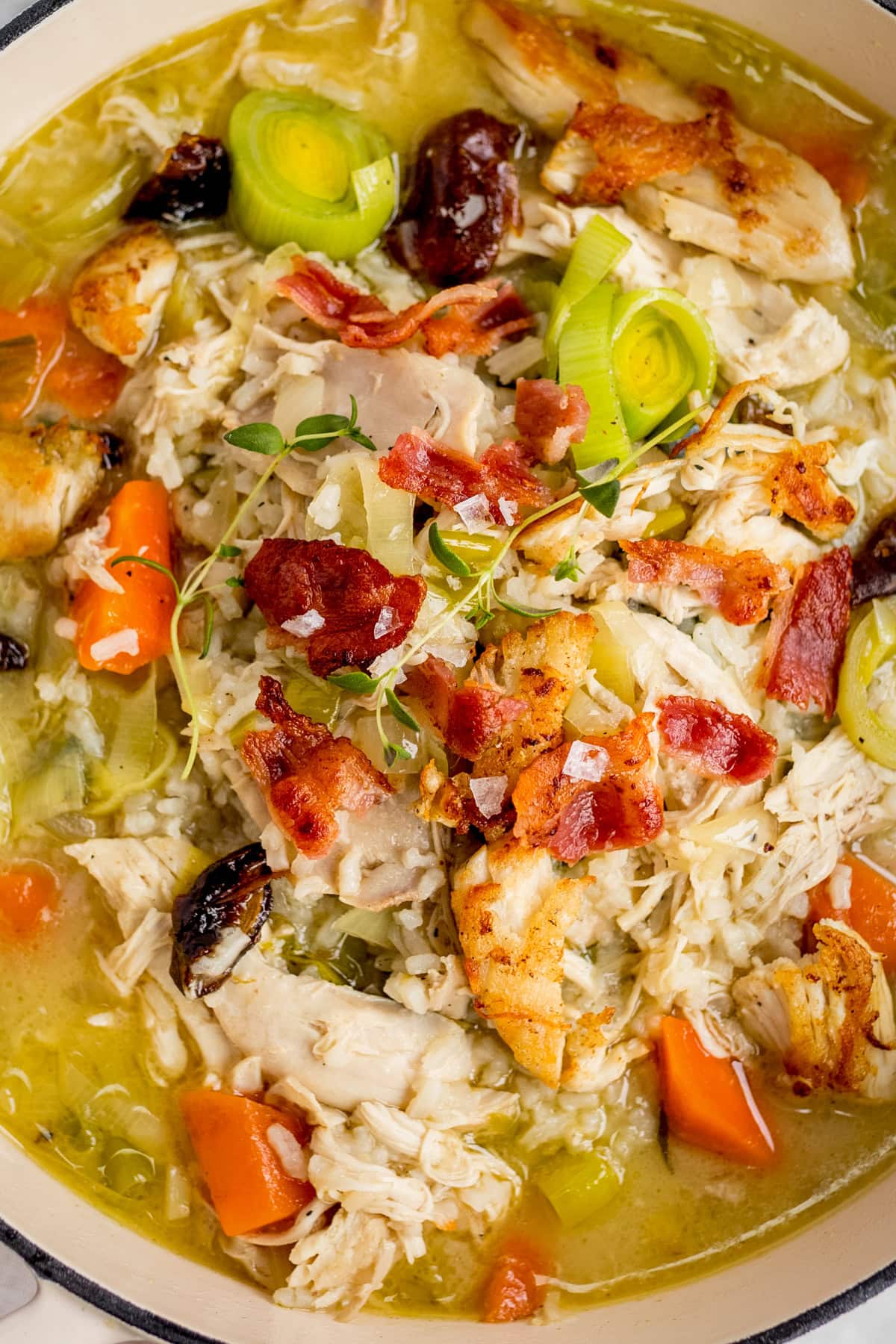 A close-up of chicken soup with shredded chicken, bacon pieces, carrots, leeks, fresh herbs, and stock in a pot.