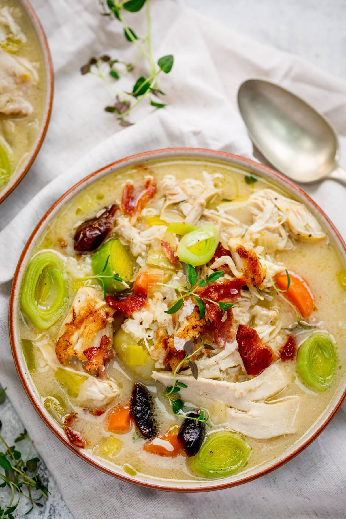 A bowl of traditional cock-a-leekie soup with shredded chicken, carrot slices, leeks, prunes, and bacon, garnished with fresh herbs, sits next to a spoon on a white cloth.