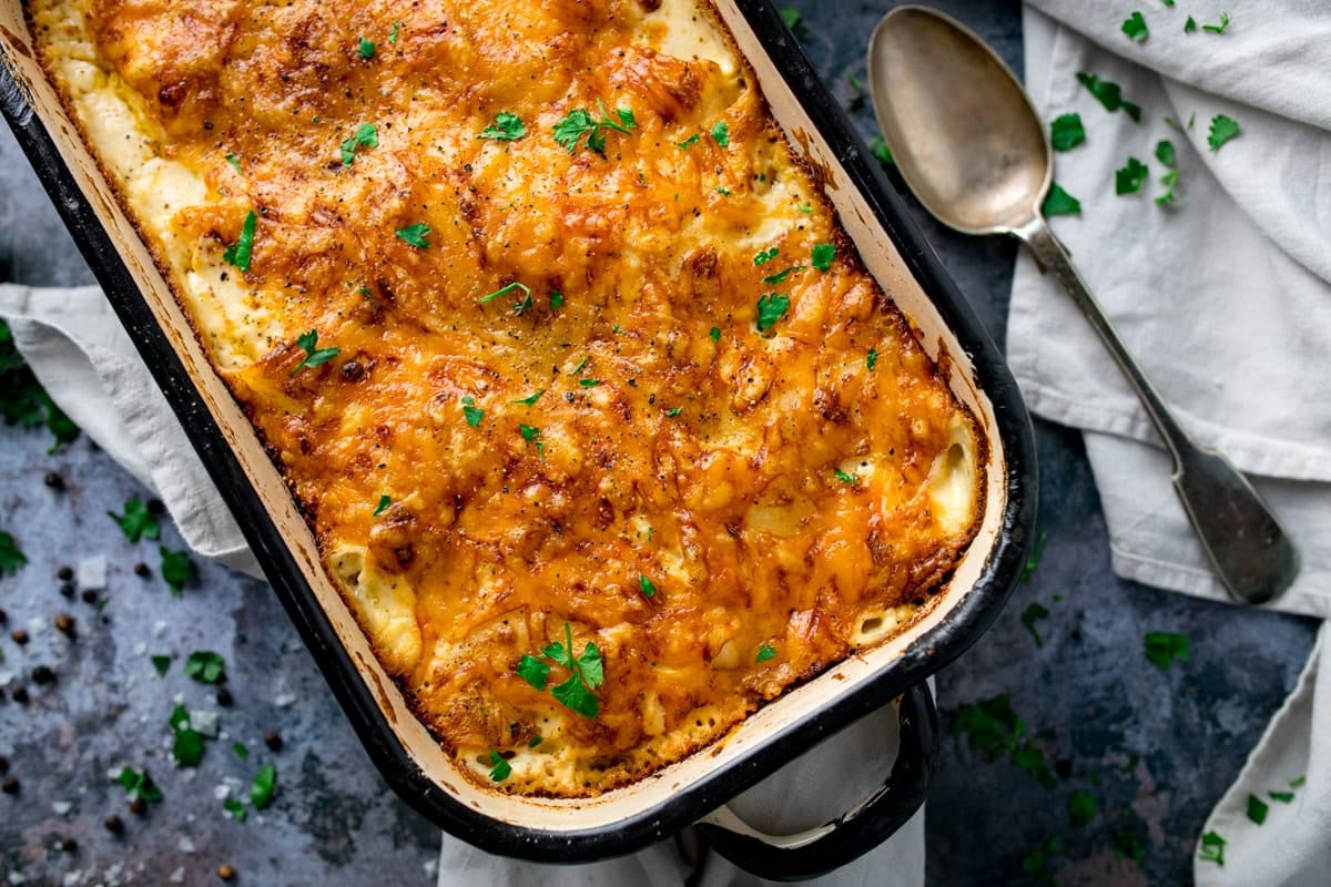 A baked casserole dish filled with cheesy, golden-brown dauphinoise potatoes, garnished with chopped parsley, sits beside a spoon and a white serviette.