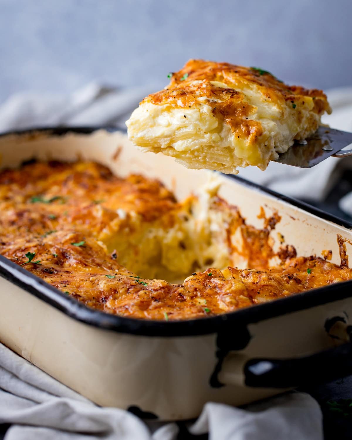 A portion of golden-brown potato dauphinoise is lifted from a rectangular baking dish, revealing creamy layers inside.