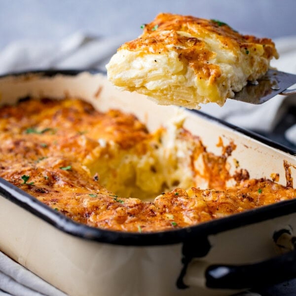 A portion of cheesy potato dauphinoise is lifted from a rectangular baking tin, showing a golden, crispy top and creamy layers within.