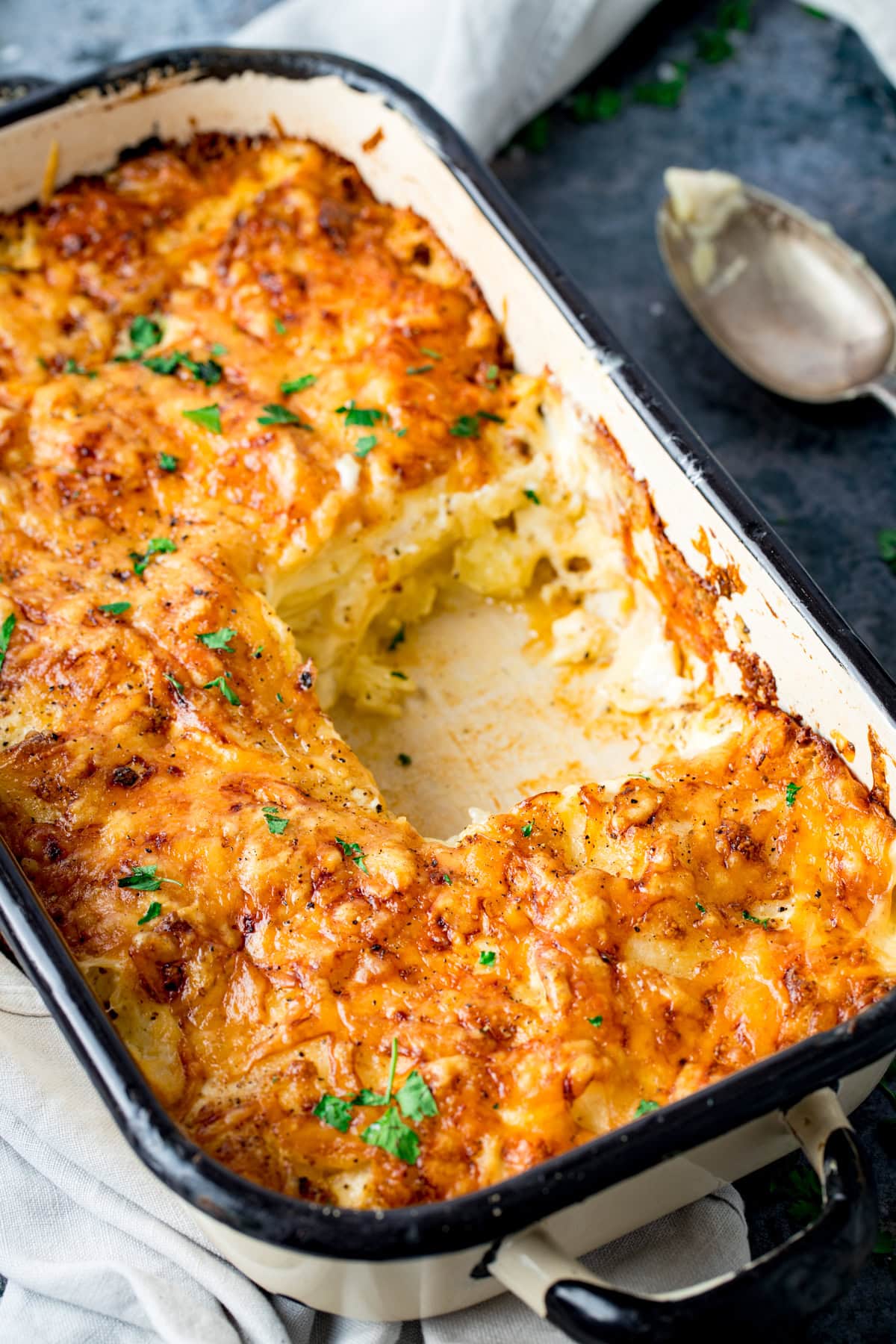 A rectangular baking dish filled with golden-brown baked cheesy potato dauphinoise, with a portion removed, revealing creamy layers inside. A spoon rests nearby on the table.