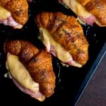 Croissant sandwiches topped with poppy seeds, each filled with melted cheese and ham, arranged on a dark baking tray.