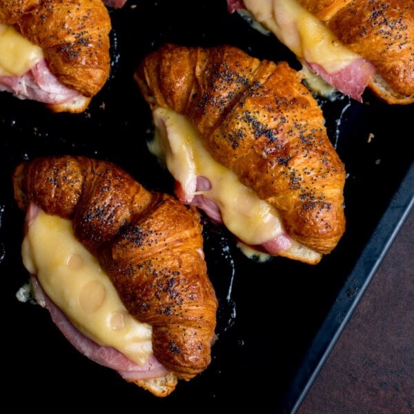 Croissant sandwiches topped with poppy seeds, each filled with melted cheese and ham, arranged on a dark baking tray.