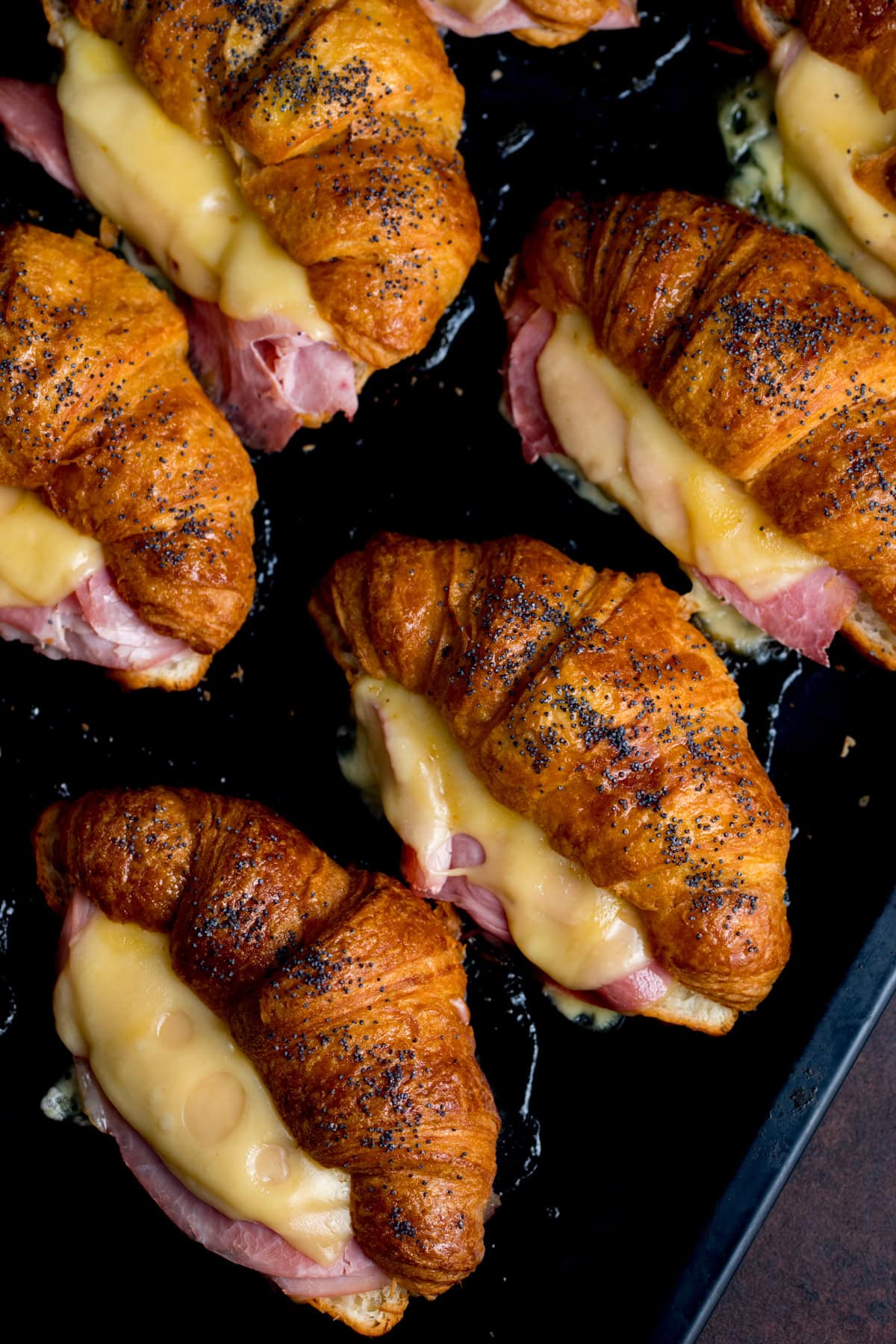 Golden filled croissants with ham and melted cheese are arranged closely on a dark baking tray.