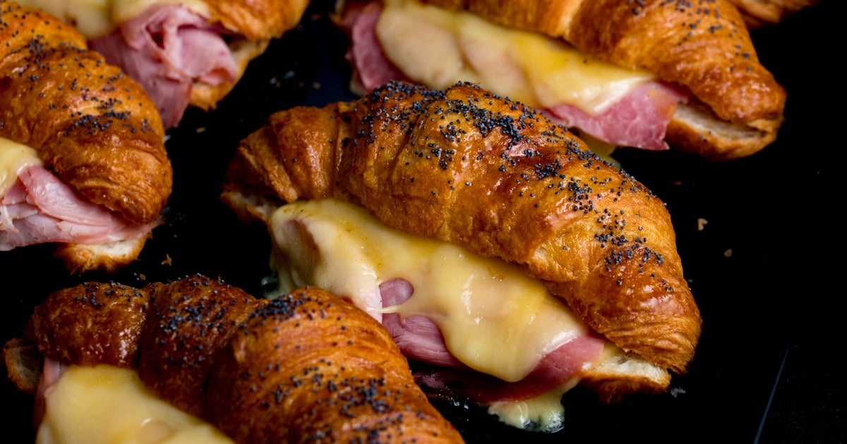 A close-up of filled ham and cheese croissants topped with poppy seeds on a dark surface.